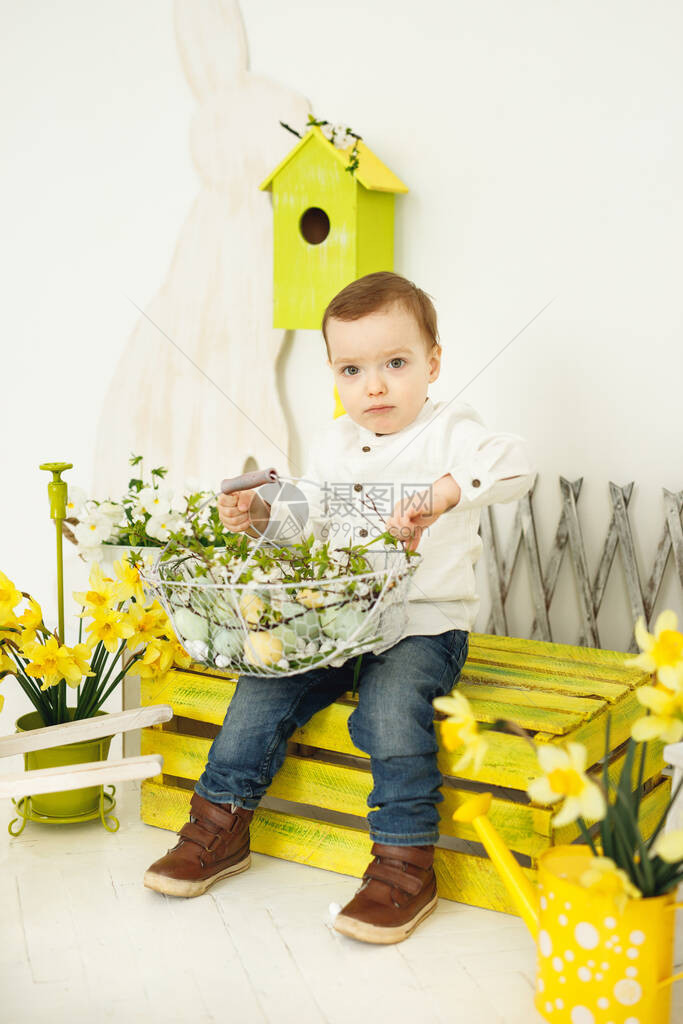 小男孩看着相机和复活节篮子的座位篮子里有五颜六色的鸡蛋的小男孩坐在黄色的长凳上概念复活图片