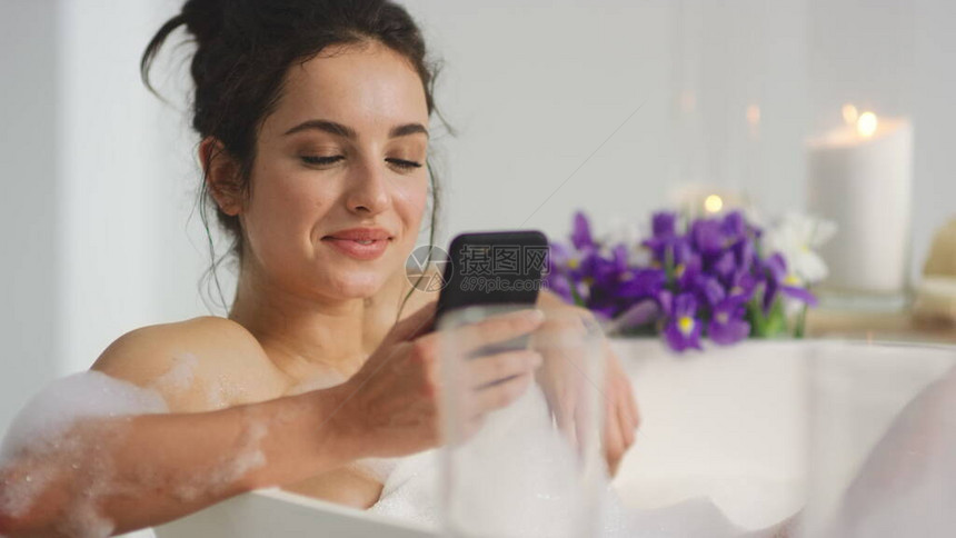 关闭了微笑的女人在慢动作浴缸里在智能手机上聊天轻松的女孩坐在浴缸里拿着手机格开朗的女人在洗澡时用图片