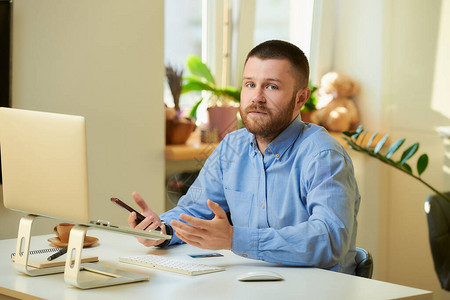 一名男子在他公寓的笔记本电脑前参加在线商务简报时被同事弄糊涂了一个穿着蓝色衬衫留着胡子的人在背景图片