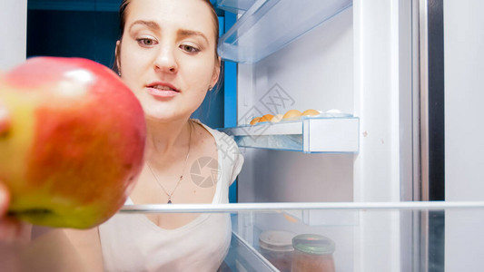 饥饿妇女晚上在厨房从冰箱里取出红苹果的肖像图片
