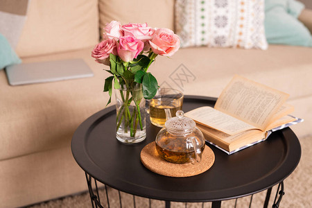 一杯水里的一束粉红玫瑰打开的书茶壶和小桌子上放着绿茶的杯子图片