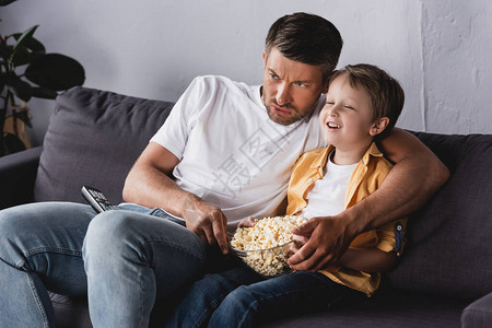担心的父亲和微笑的儿子在家看电视和在沙图片