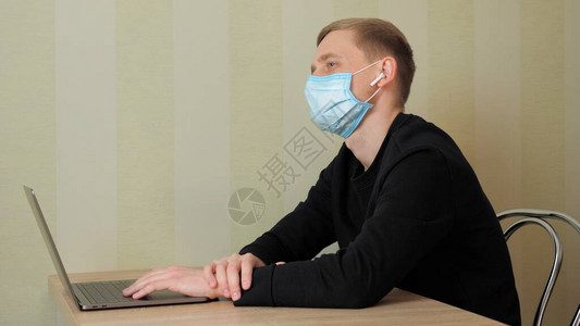 戴着防护医用口罩和耳朵里的无线耳机的男人在家里用笔记本电脑工作图片