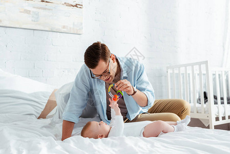快乐的年轻男人玩着婴儿响铃在床上躺着可图片