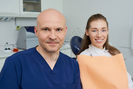 一位光头的中年牙医和一位面带微笑的漂亮女病人正在牙医的办公室里摆姿势图片