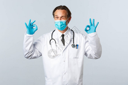 戴着医用口罩和手套的开朗热情的医生表现出良好的姿态和眨眼图片