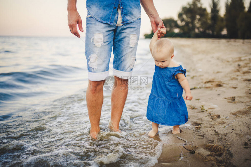 父亲和女儿手牵着手女蹒跚学步的女婴在日落时分在海边的沙滩上牵着父亲的手一起旅行爱家庭父亲节假期概念爸图片