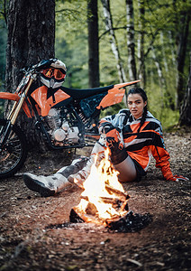 美丽的年轻女赛车手穿着摩托穿梭装坐在营火图片