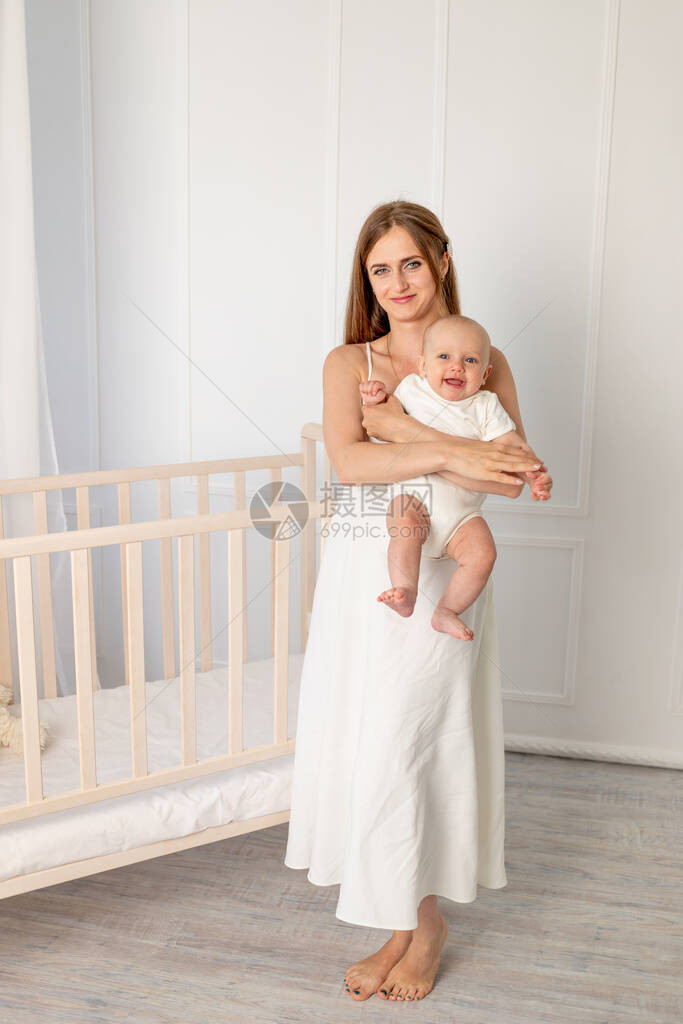 年轻漂亮的母亲抱着她6个月的女儿在婴儿床旁站在婴儿床旁图片