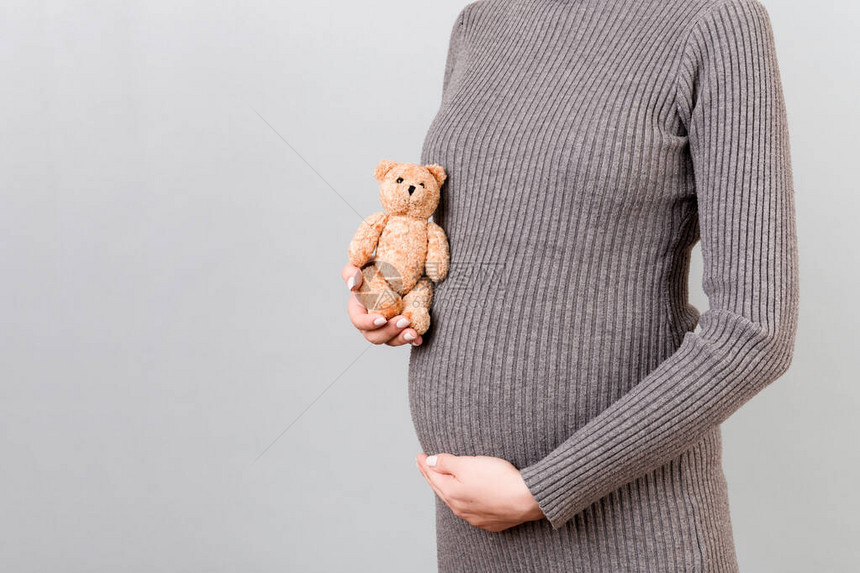 身穿灰色连衣裙的孕妇在灰色背景下抱着一只泰迪熊靠在肚子上的特写年轻的母亲正在怀图片