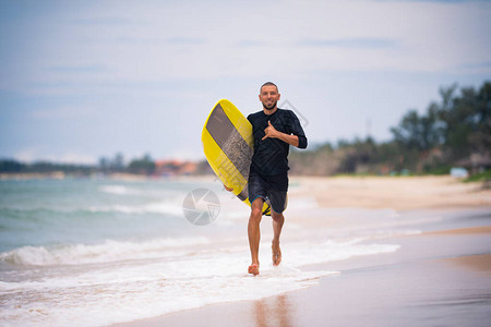 穿着黄色冲浪板长的健身冲浪员在阳光明媚的海滩上跑动图片