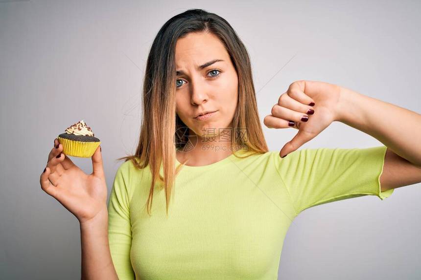 青蓝眼睛的年轻美女在白色背景上吃甜巧克力蛋糕脸发怒负表情示不喜欢用拇指往下看拒图片
