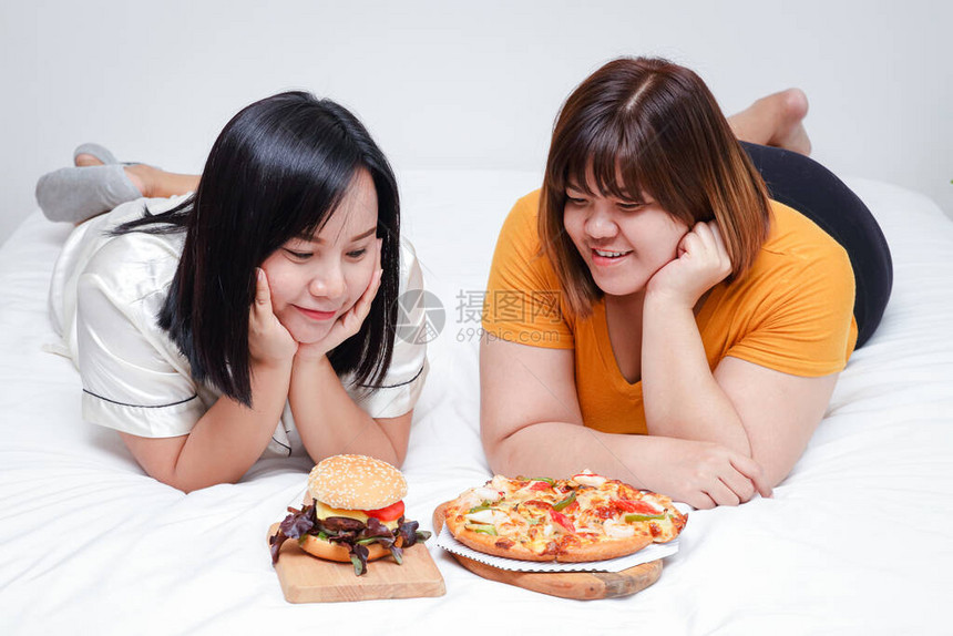 两个胖女孩在卧室的床上吃披萨和汉堡图片