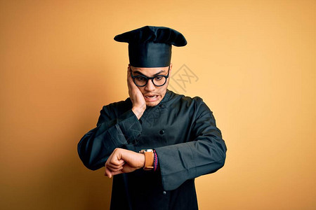 身穿炊具制服和帽子的巴西年轻厨师在孤立的黄色背景中看着手表时间担图片
