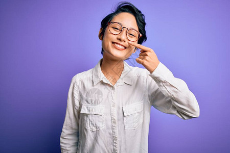年轻漂亮的亚洲女孩穿着休闲衬衫和眼镜站在紫色背景上图片