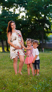新生命概念长棕色头发的孕妇和两个小孩在公园草坪上摆布小女孩摸母亲腹图片