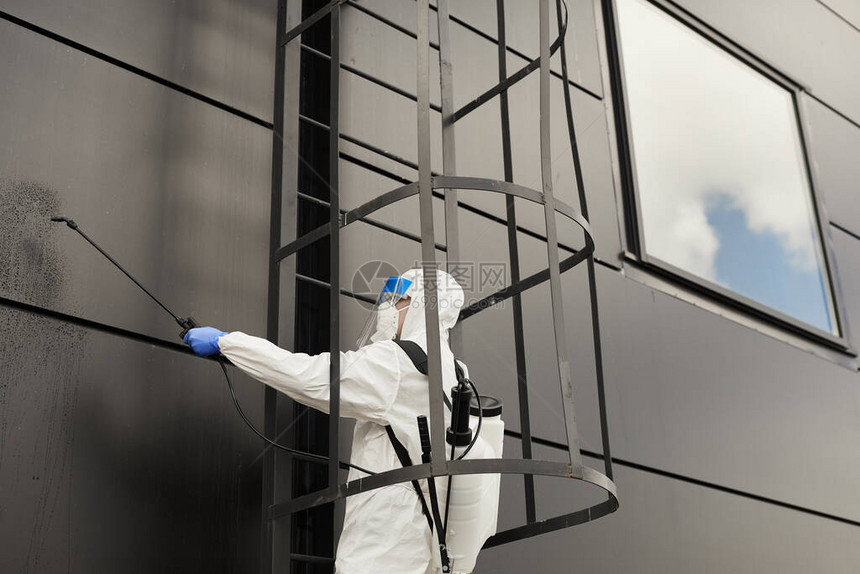 一名身着防护服的工人在消毒或清洁复制空间期在黑建筑外墙上喷洒化学品图片