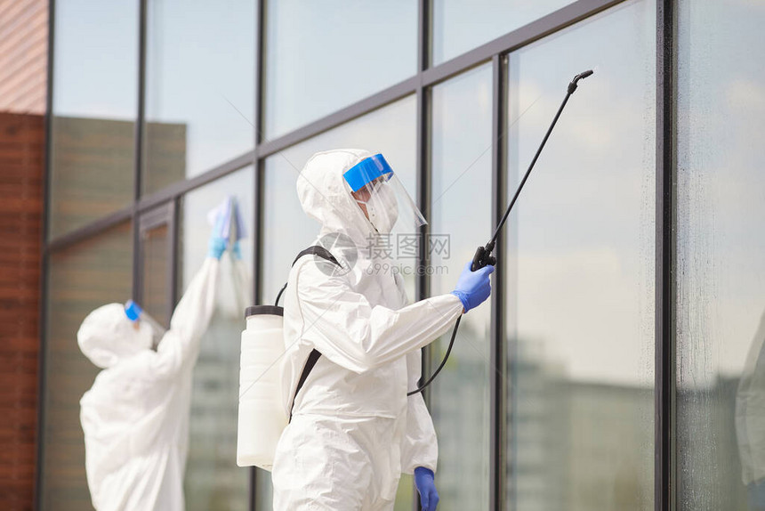 两名身穿防护服的工人在消毒或清洁期间在户外建筑物上喷洒化学物质的画图片
