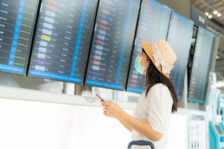 戴面罩的亚洲女游客从机场候机楼到达出发板检查航班图片