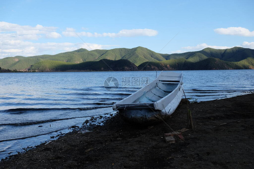 一条白色的小船搁浅在黑土海滩上蓝色的湖水在四周荡漾青山背景晴朗的蓝天在中图片