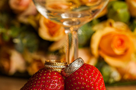 新娘和新郎的金色结婚戒指放在玻璃杯和一束玫瑰花前的草莓上图片