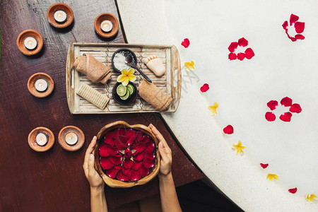 女手握着红玫瑰花瓣的木碗带有石灰洗涤剂盐毛巾和木盘刷子的罗马式温泉图片