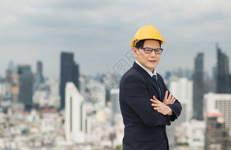 年轻英俊的亚洲土木工程师站在建筑城市背景的商业套房中身着头盔黄色硬帽图片