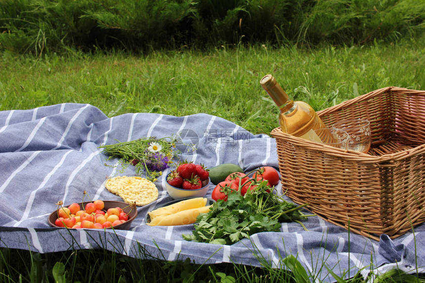 夏季野餐食品一瓶白葡萄酒玻璃杯草莓碗里的樱桃新鲜蔬菜西红柿欧芹鳄梨灰色和白色条纹毯子上的图片