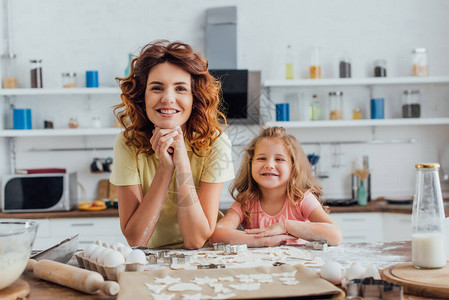 年轻母亲和女儿在厨房桌上的生饼干和原料旁看照相图片