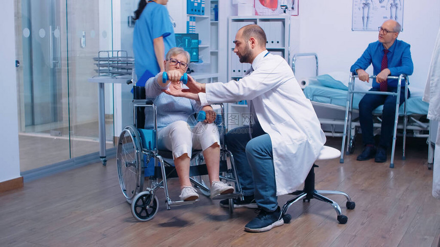 医生在一家私人康复诊所帮助坐在轮椅上的老年残疾妇女恢复肌肉力量使用哑铃锻炼的无效的人康复医院图片