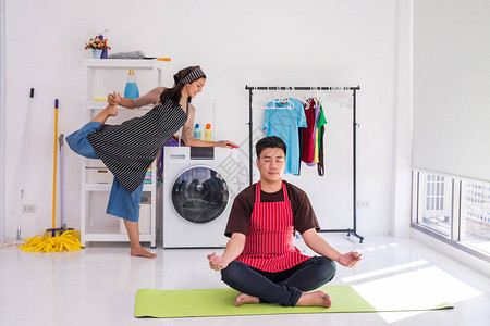 快乐的年轻亚洲夫妻和围裙一起在等待洗衣机洗衣服时一起做瑜伽周末做家图片