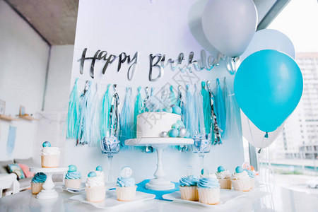 庆祝一岁男婴的节日装饰架子上的白蛋糕纸杯蛋糕蓝色球和金属丝题字背景图片