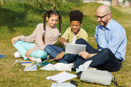 微笑的男教师坐在绿草地上与儿童交谈在阳光下享受户外课的完整肖图片