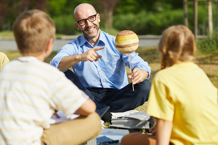 光天化日照复制空间在户外课与一群儿童交谈时微笑和指着行星模型的秃图片