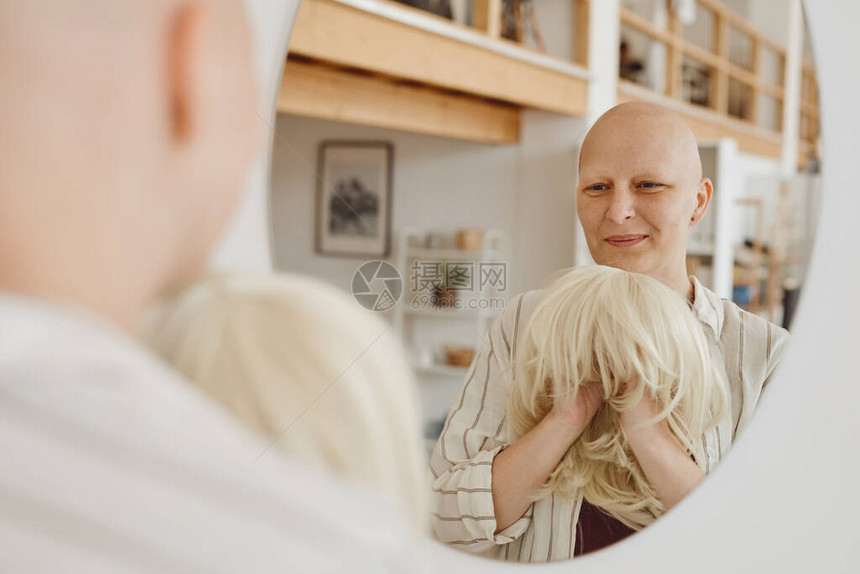 秃头成年妇女看着照镜子戴着假发站在温暖的室内高视和癌症意识复制空图片