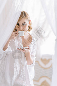一个穿着白衬衫的年轻金发美女在轻便浴室享受早上咖啡柔软的选择聚焦图片