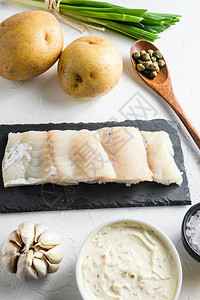 传统的鱼和薯条配料方在石板面糊土豆鞑靼酱柠檬刺山柑绿色香草大蒜盐胡椒粒上的生鳕鱼片在白石背景侧视图片