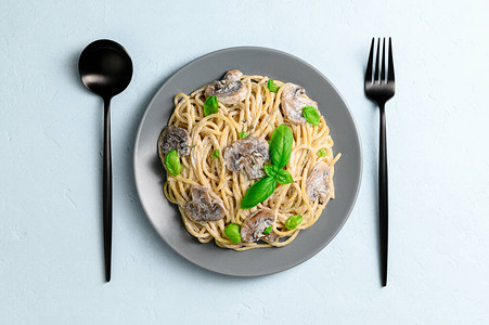 意大利面配奶油酱蘑菇和浅蓝色背景上的绿色罗勒用黑色勺子和叉子放在灰色盘子里的意大利背景图片
