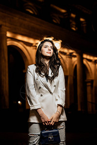 有魅力的女人穿着白色时尚夹克和牛仔裤的垂直照片抬头有照明背景的建筑图片