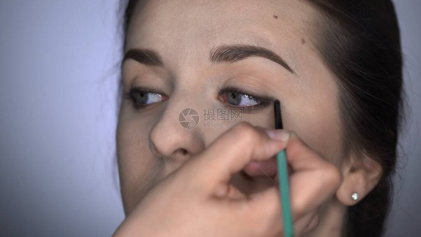 坐在工作室的美丽而华丽的女人的专业化妆过程化妆师在棕色眼睛女孩的上眼睑上涂抹眼线图片