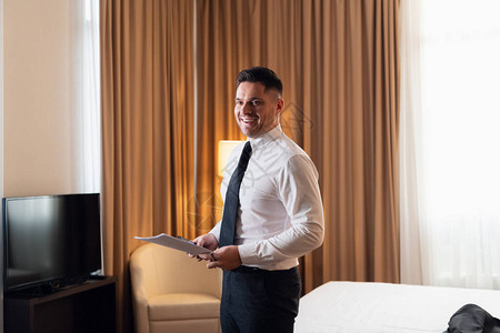 一位年轻快乐的旅行者商人穿着一件白衬衫和一条领带站在旅馆房间里图片