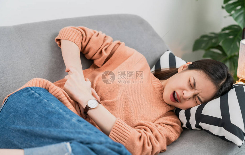 年轻的亚裔女手握着沙发上的胃部和疼痛期的抽筋图片
