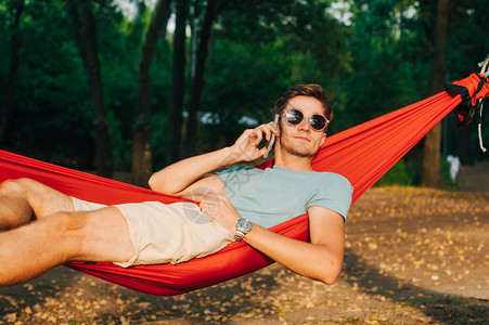 一个戴着墨镜和休闲服的年轻人躺在公园的吊床上图片