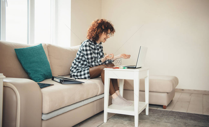 一位有卷头发和眼镜的caucasian女士在家中使用笔记本电脑上网课时笑着微图片