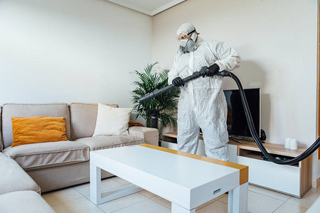 穿着PPE的男子用COVID19消毒机对房子的客厅进行消毒大流行背景图片