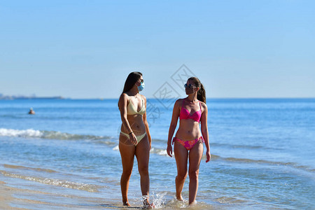 两名身戴面罩和比基尼的年轻妇女在靠近海边走路时相互交谈图片
