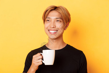 穿着金色头发的英俊年轻亚洲人看着和微笑喝着咖啡或茶站在黄色背图片