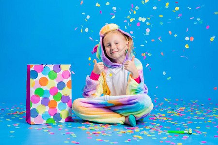 一个穿着独角兽制服的快乐小女孩拿着一个装有彩色纸屑的礼品袋生日假期或销售的概背景图片