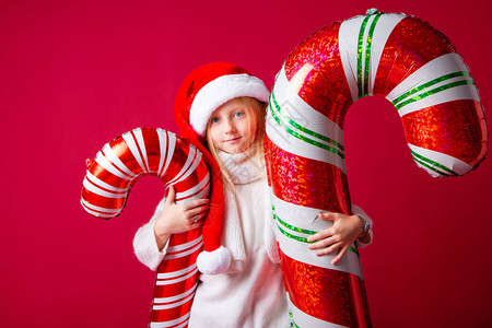一个戴着圣诞老人帽子的小女孩拿着红背景的糖果甘蔗形气球圣诞节的概图片