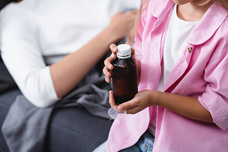 儿童在患病母亲附近持有一瓶糖浆和勺子的图片